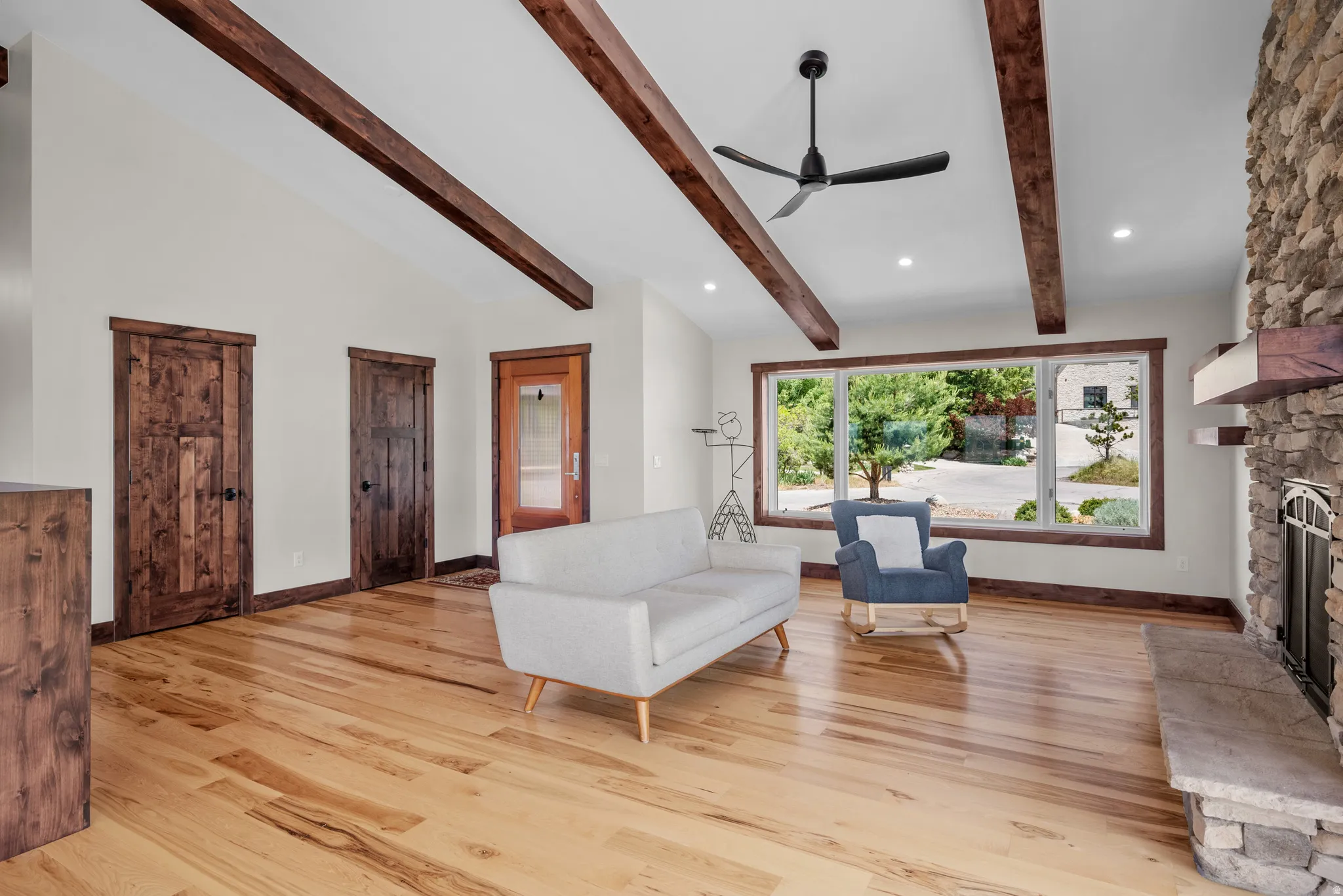 Vaulted ceilings featuring exposed wood beams, a stone-clad fireplace with a stone hearth, expansive windows with wood trim, wood-finish flooring, and dark wood paneled doors