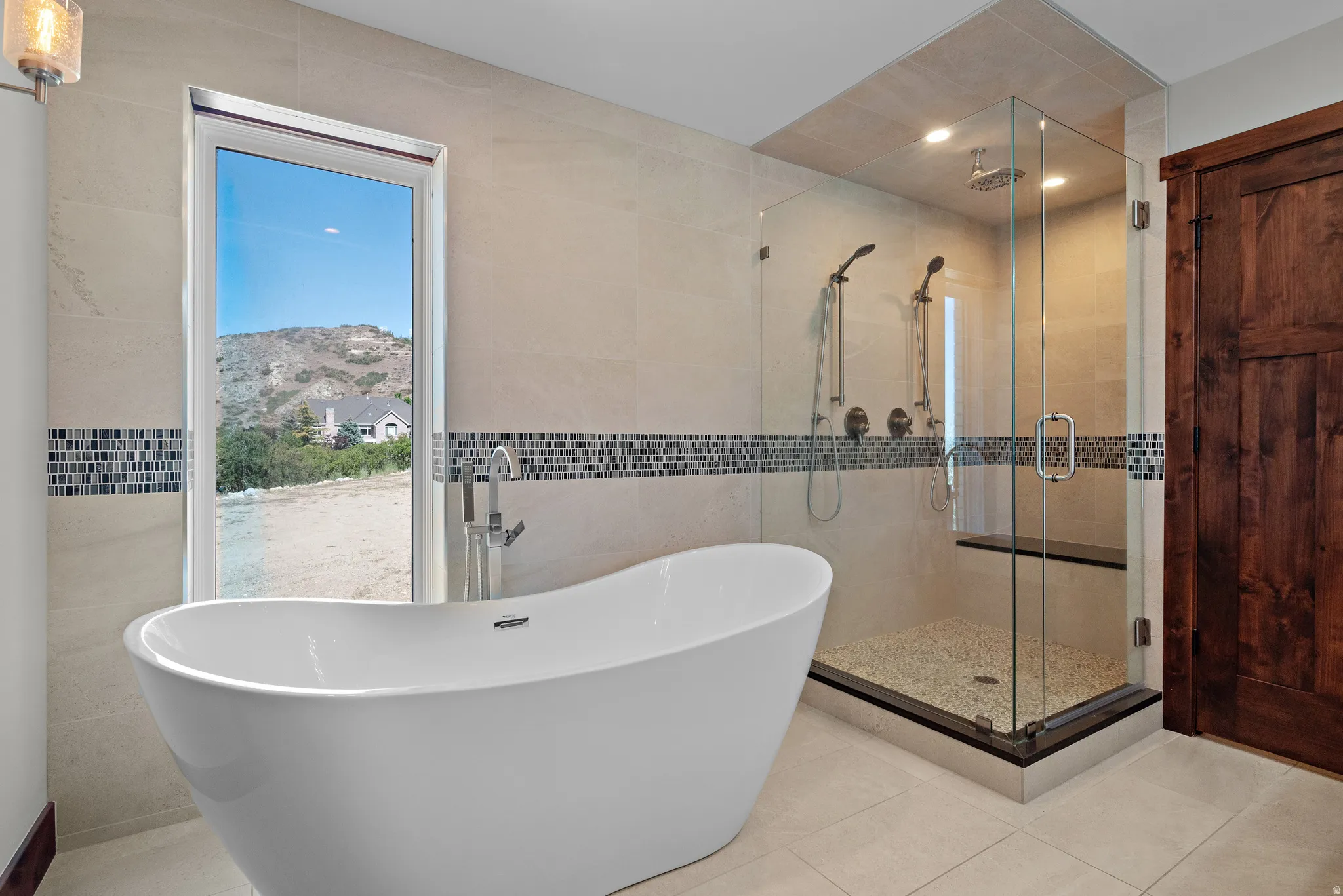 Modern bathroom featuring a freestanding soaking tub with floor-mounted faucet, a glass-enclosed shower with dual shower heads and a built-in bench, light-toned tile walls with a mosaic accent band, and a rustic wood door