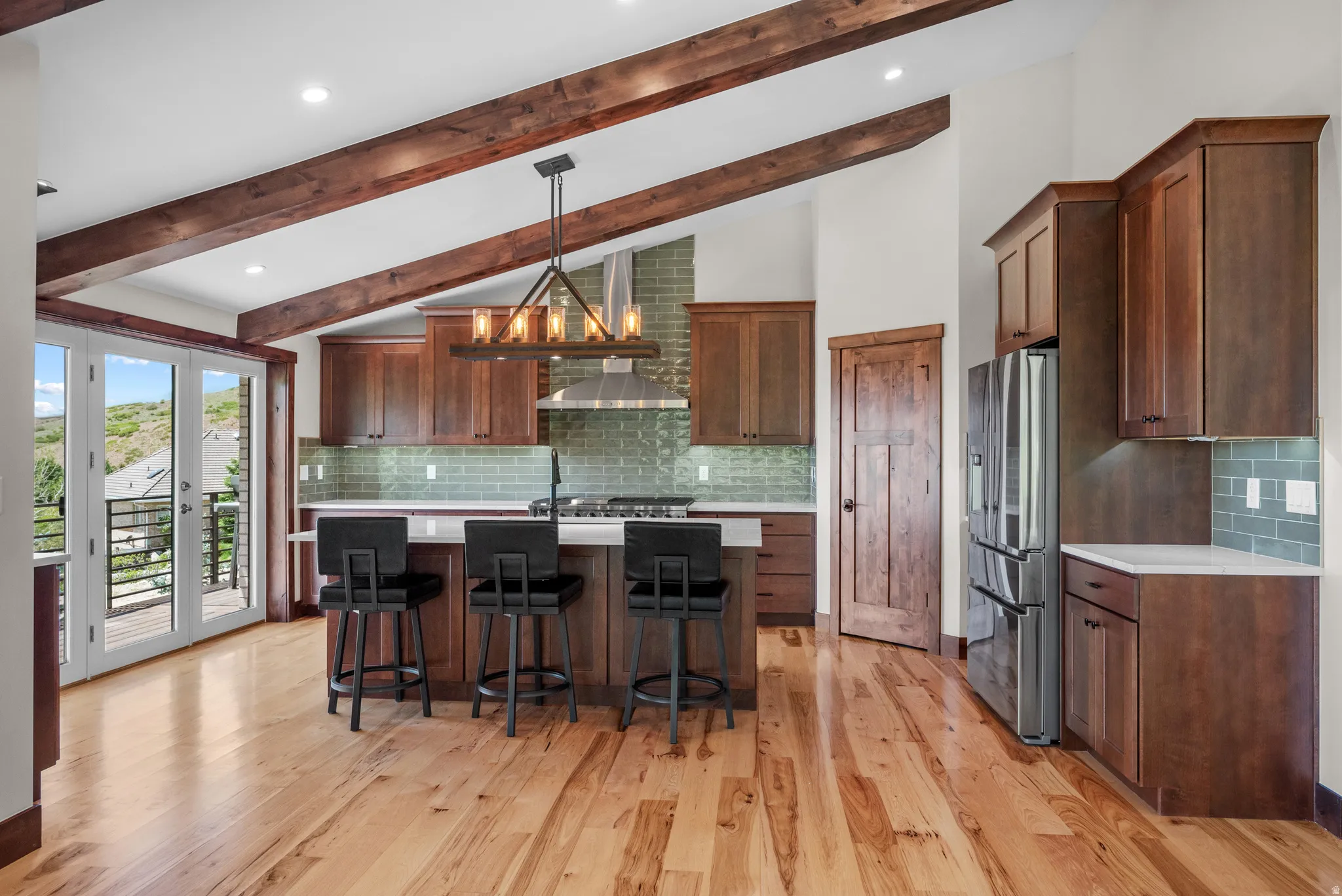 Spacious kitchen featuring exposed wood ceiling beams, wood-finish flooring, a central island with countertop seating, stainless steel appliances, and French doors opening to a balcony