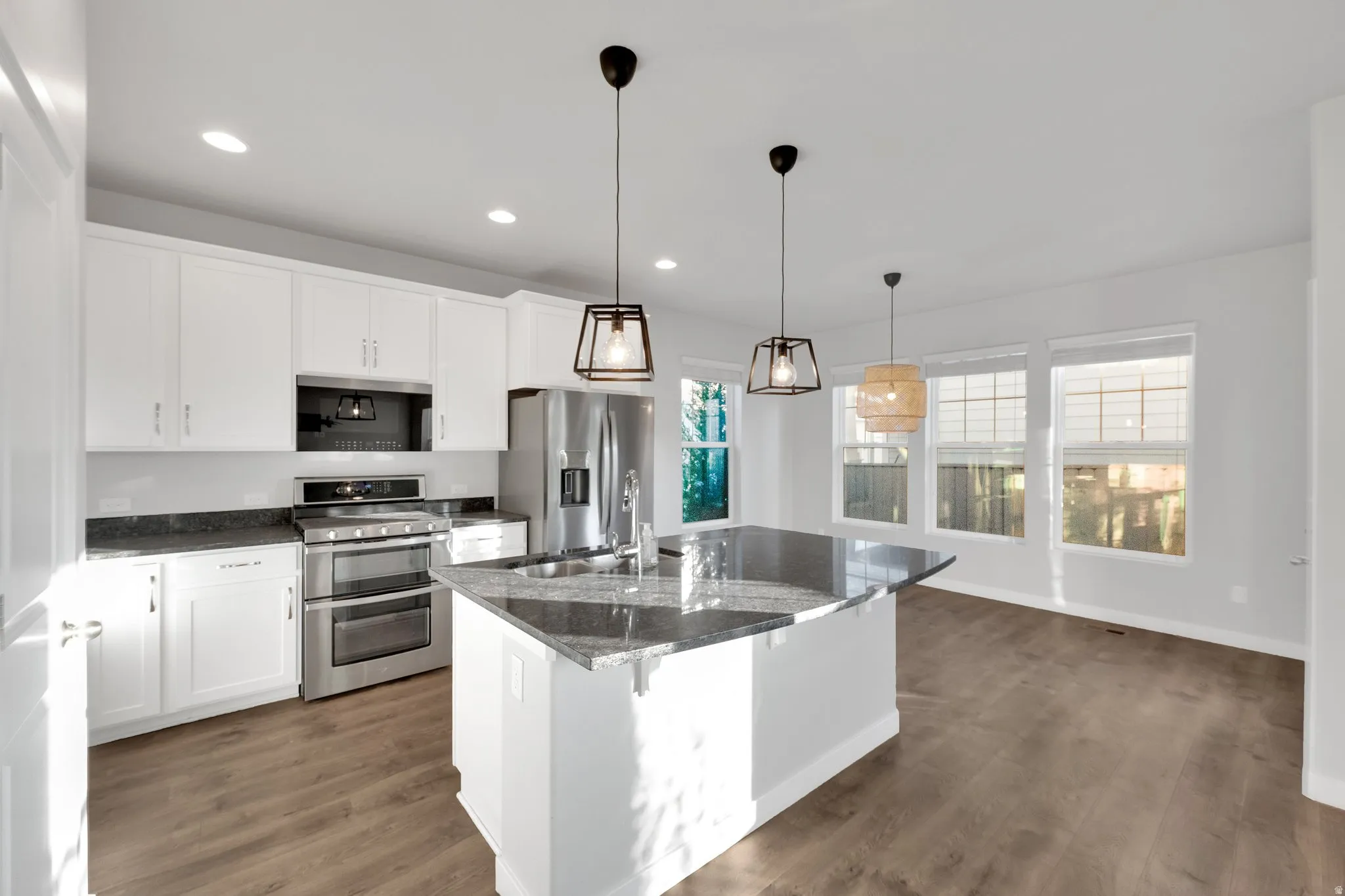 Contemporary kitchen featuring white cabinetry, stainless steel appliances, dark stone countertops, a central island with an undermount sink, and wood-finish flooring
