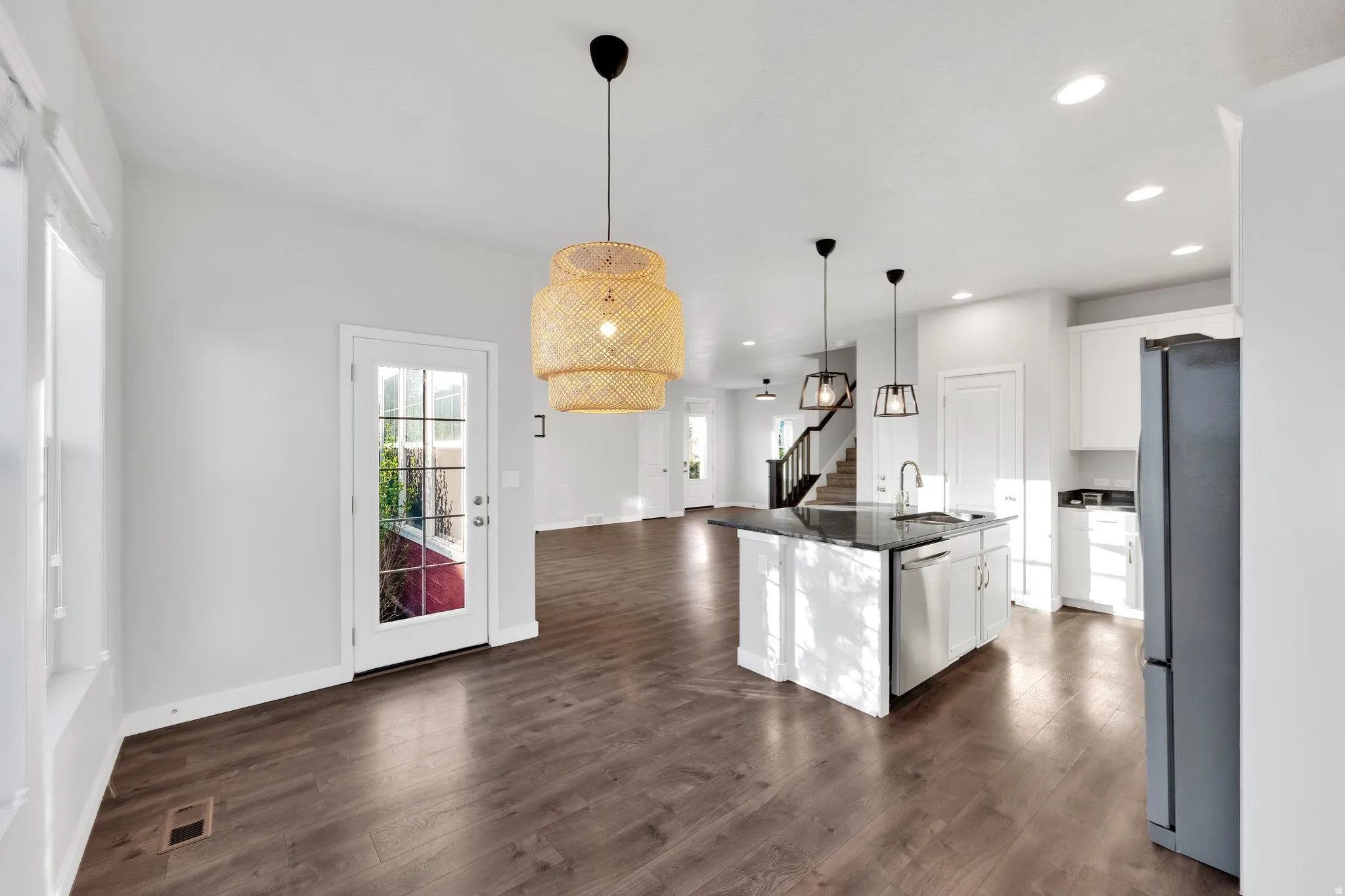 Open-concept kitchen and dining area featuring dark wood-finish flooring, a white kitchen island with a dark countertop, stainless steel appliances, and multiple decorative light fixtures