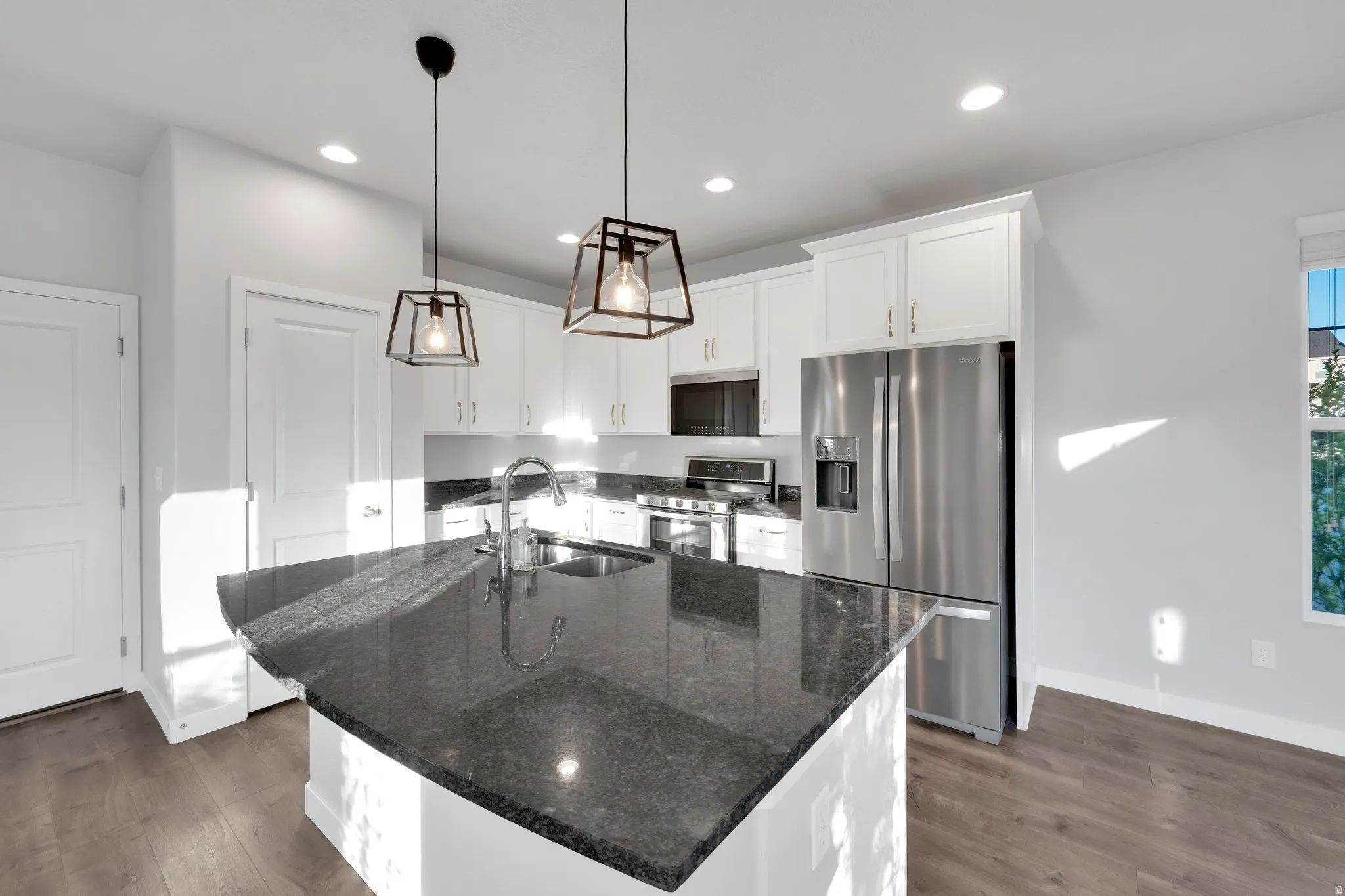 Kitchen island with a dark countertop, stainless steel appliances, white cabinetry, wood-finish flooring, and recessed lighting