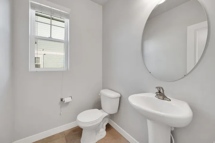 Powder room featuring a pedestal sink with chrome faucet, a window with blinds, a toilet, an oval mirror, and tile flooring