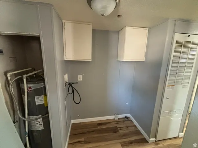 Utility area featuring wood-finish flooring, two upper wall cabinets, a wall-mounted furnace, and a water heater in an alcove