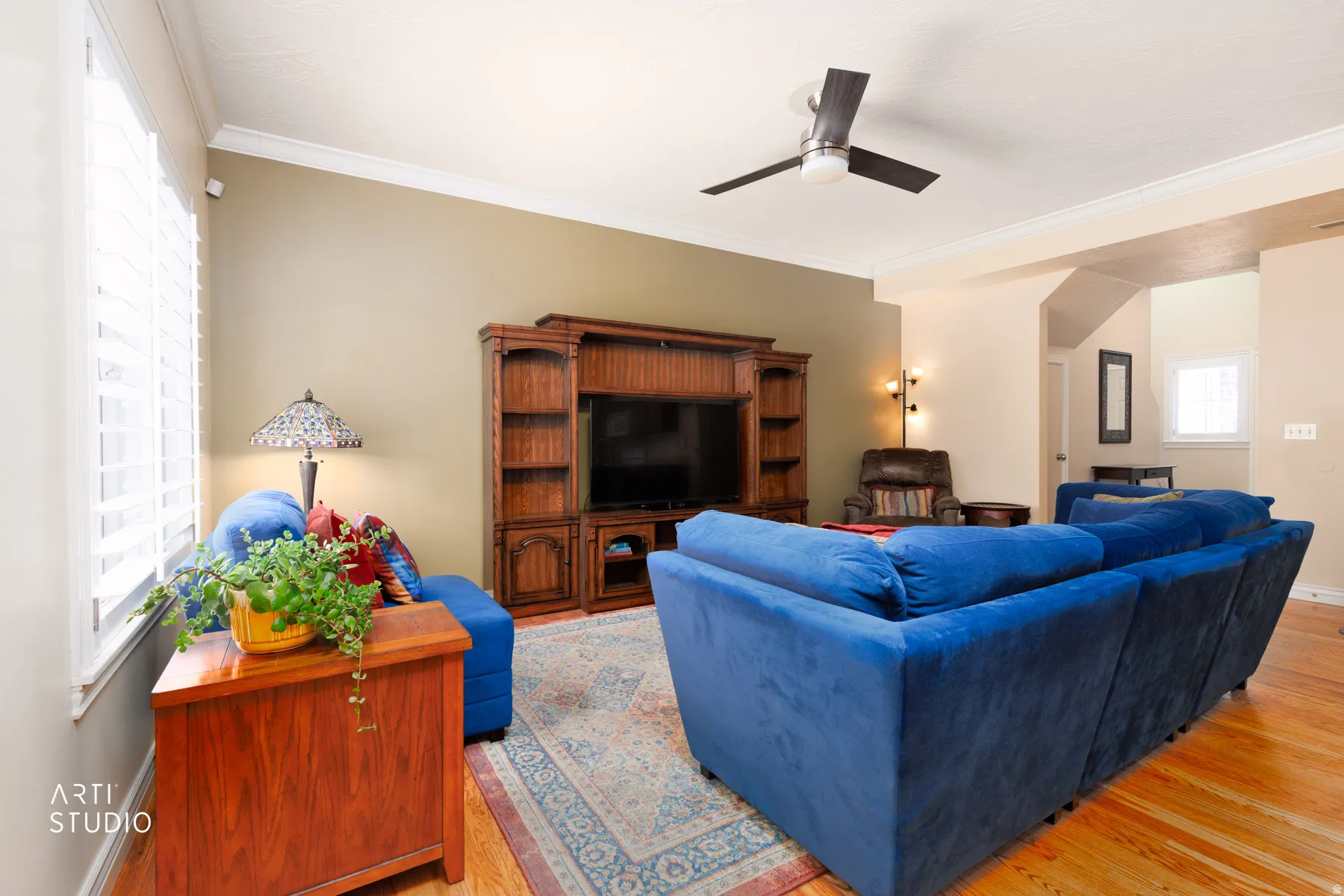 Living area featuring wood-finish flooring, a built-in entertainment center with shelving, crown molding, and a ceiling fan with integrated lighting