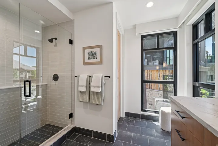 Glass-enclosed shower with subway tile surround and dark-finish fixtures, complemented by dark-framed windows and wood-finish cabinetry with a light-toned countertop