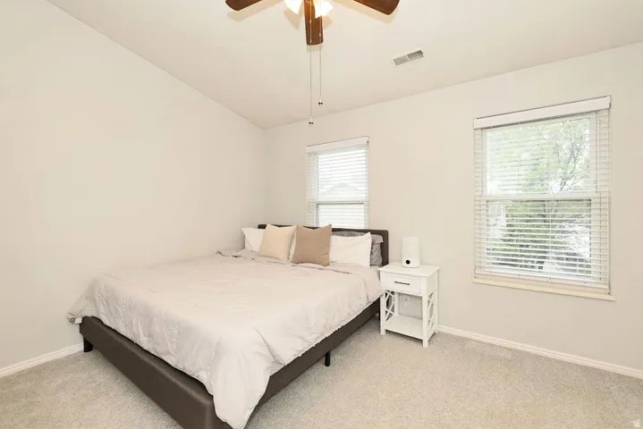 Bedroom featuring light-colored carpet flooring, two windows with blinds, a ceiling fan with integrated lighting, and neutral wall paint