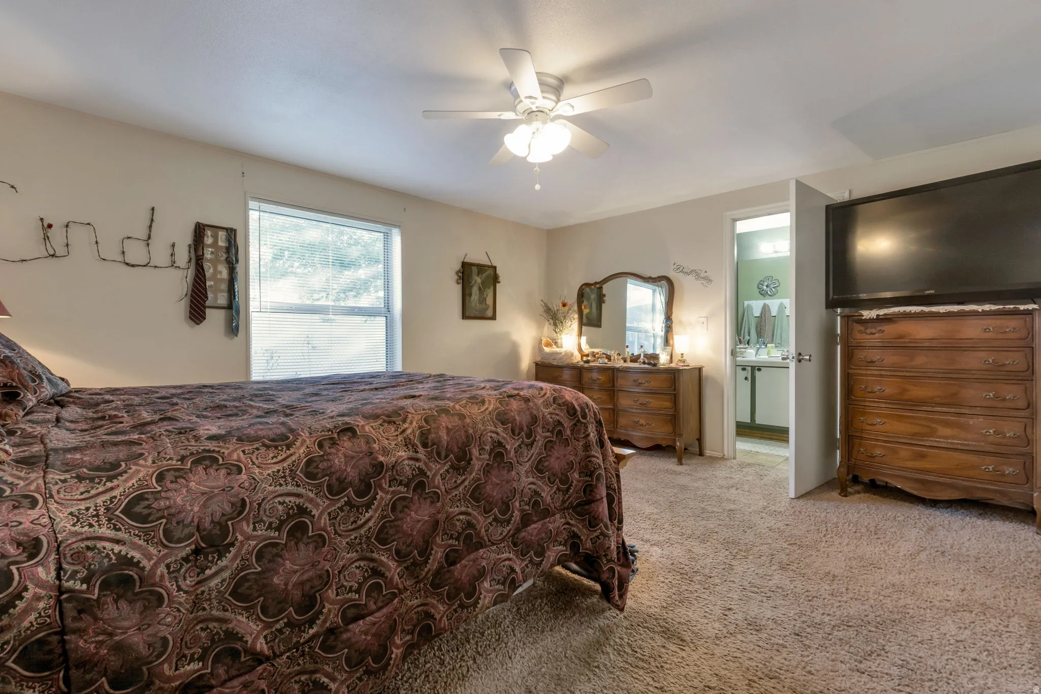 Carpeted bedroom featuring a ceiling fan with integrated lighting, a window, and a door opening to a bathroom with a vanity and mirror