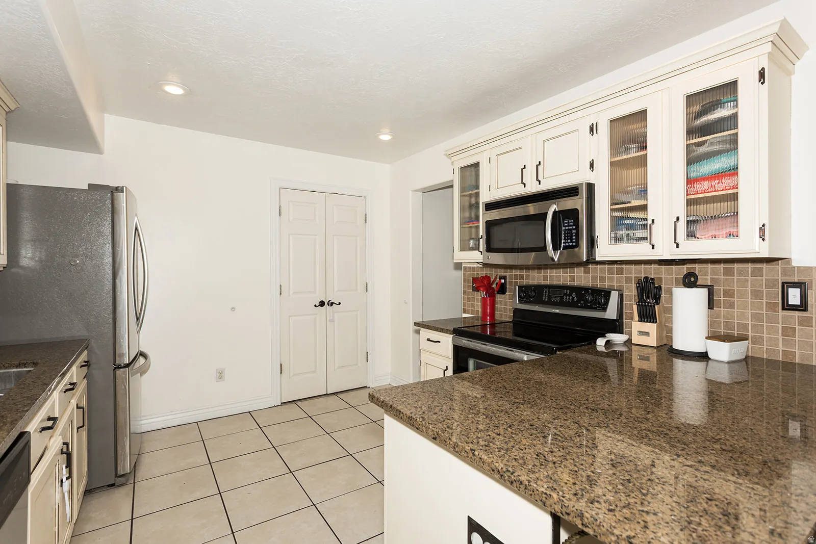 Kitchen featuring light-toned cabinetry with glass-front uppers, stainless steel appliances, speckled countertops, a tiled backsplash, and square floor tiles