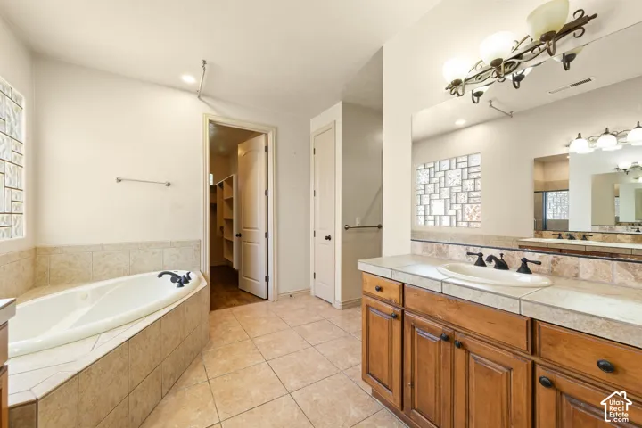 Bathroom featuring a corner soaking tub with tile surround, a double vanity with wood cabinetry and tile countertop, and a glass block window