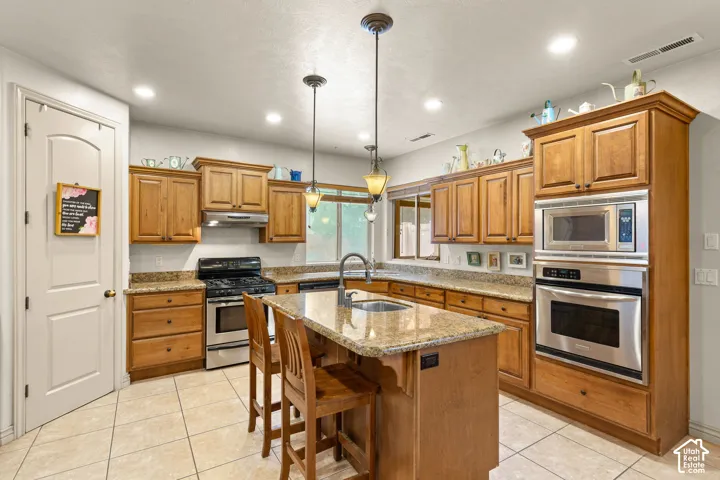Spacious kitchen featuring warm wood cabinetry, granite countertops, stainless steel appliances, a center island with sink, and tile flooring
