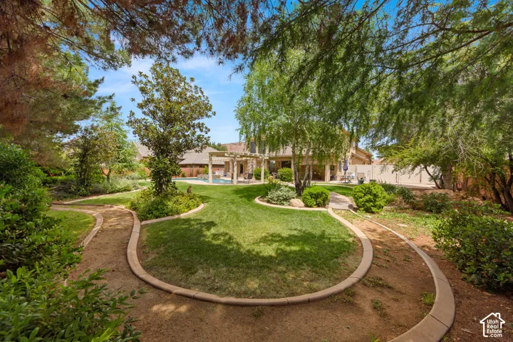Expansive backyard featuring a lush green lawn, mature trees, winding garden paths, a distant pool, and a shaded patio structure