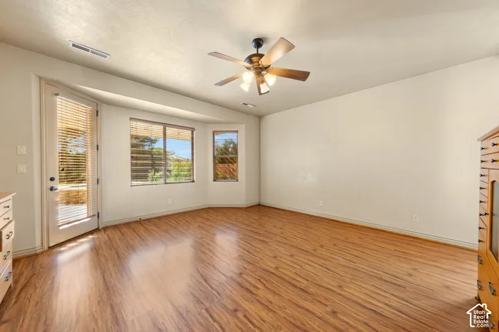 Spacious room featuring wood-finish flooring, a ceiling fan with integrated lighting, and multiple windows with horizontal blinds