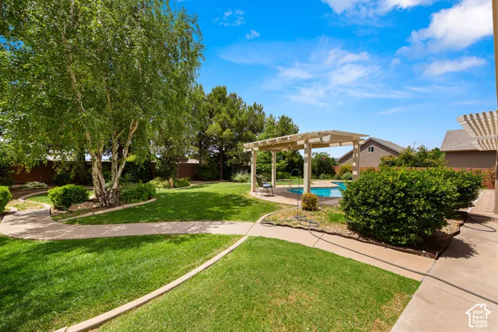Spacious backyard featuring a swimming pool, a wood pergola, lush green lawn, mature trees, and concrete pathways
