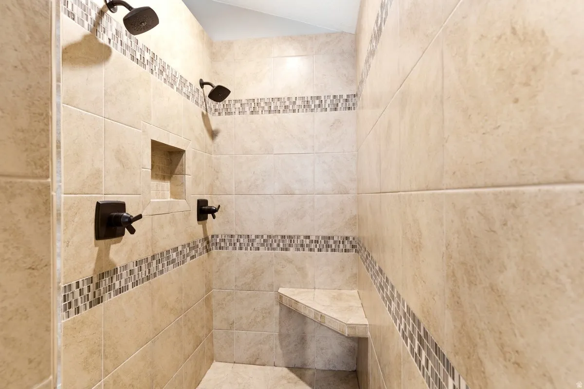 Tiled shower featuring dual shower heads, a built-in niche, a corner bench, and decorative mosaic tile accents