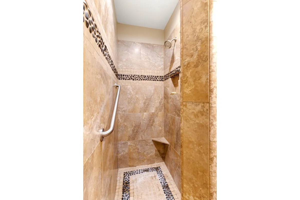 Walk-in shower featuring natural stone-look wall tiles, pebble-detail accent bands, a built-in shower bench, and a brushed nickel grab bar