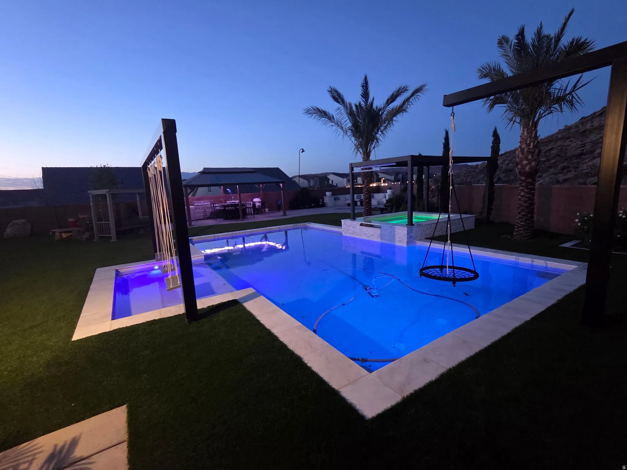 Illuminated swimming pool with integrated spa and waterfall feature, surrounded by artificial turf and concrete pavers