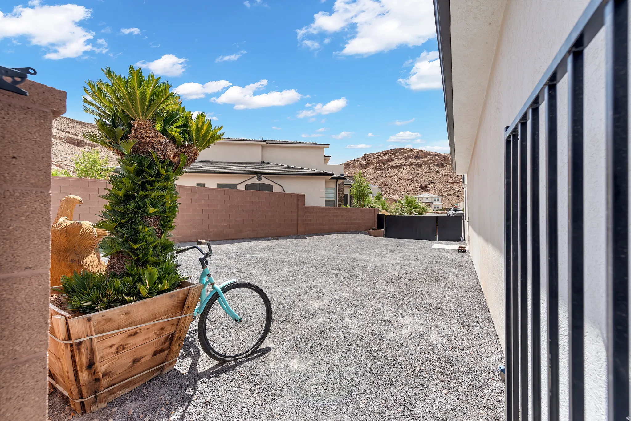 Gravel-covered outdoor space featuring a stucco exterior with black metal fencing, a large wood planter, and a mature palm plant