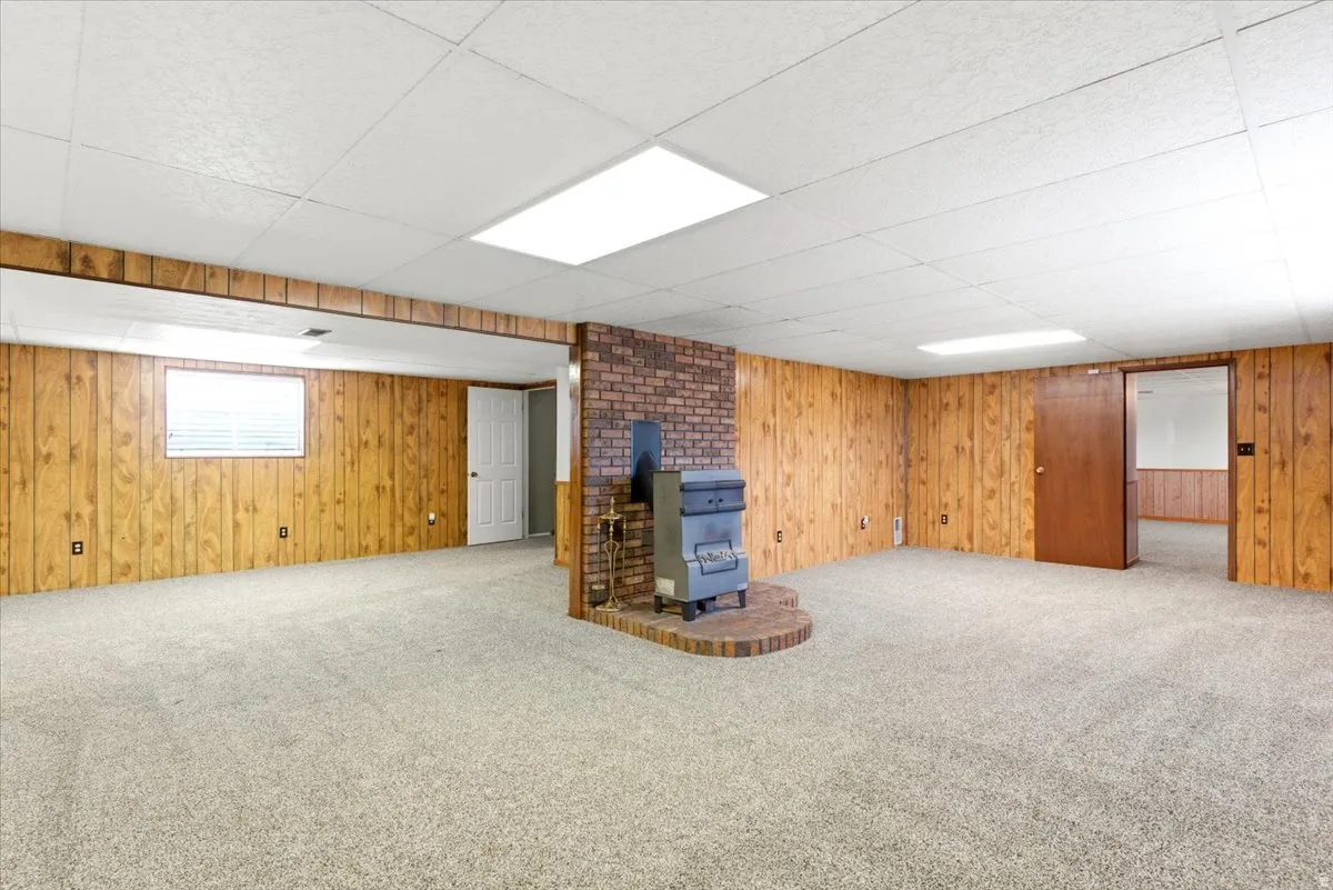 Spacious lower-level room featuring wood paneling, light-colored carpeting, a brick fireplace surround with a wood-burning stove, and a drop ceiling with recessed lighting