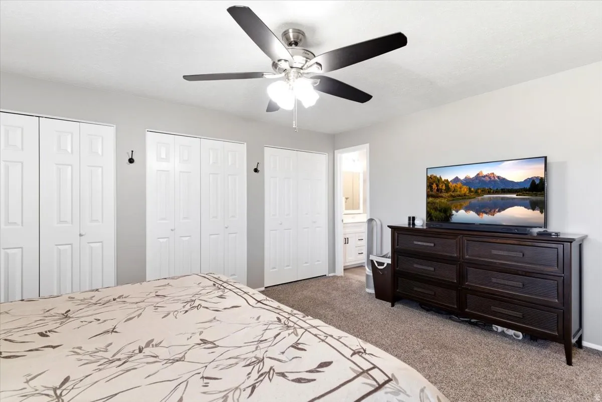 Carpeted room featuring multiple bifold closet doors, a ceiling fan with integrated lighting, and direct access to a bathroom with a vanity and mirror