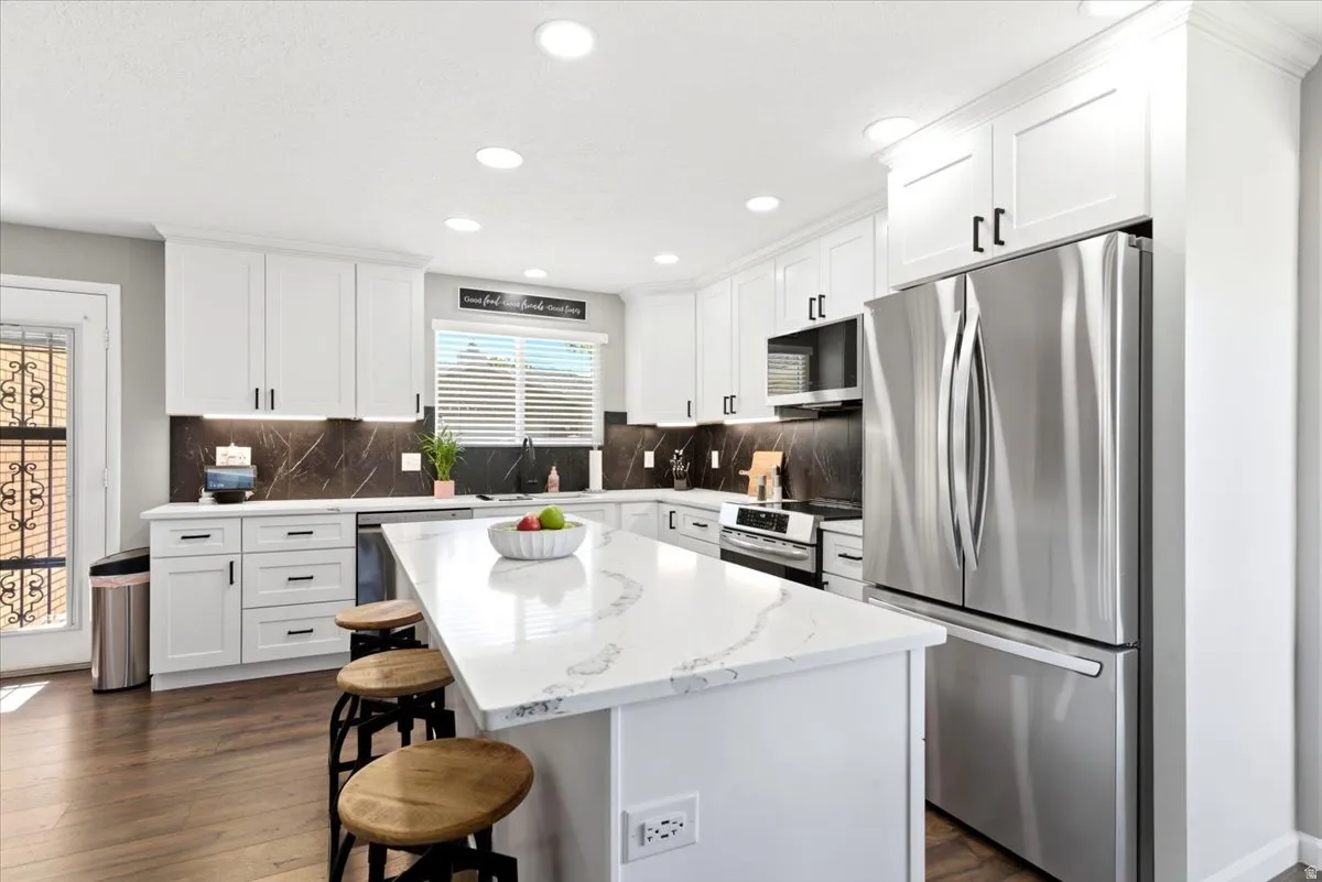 Modern kitchen featuring a large central island with a waterfall-edge countertop, white shaker-style cabinetry, stainless steel appliances, and wood-finish flooring
