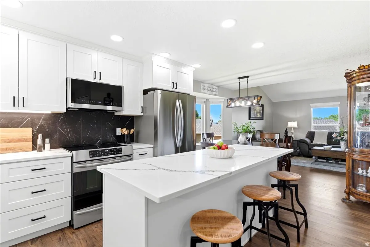 Open-concept kitchen featuring white cabinetry, stainless steel appliances, a black marble-patterned backsplash, and a large central island with seating