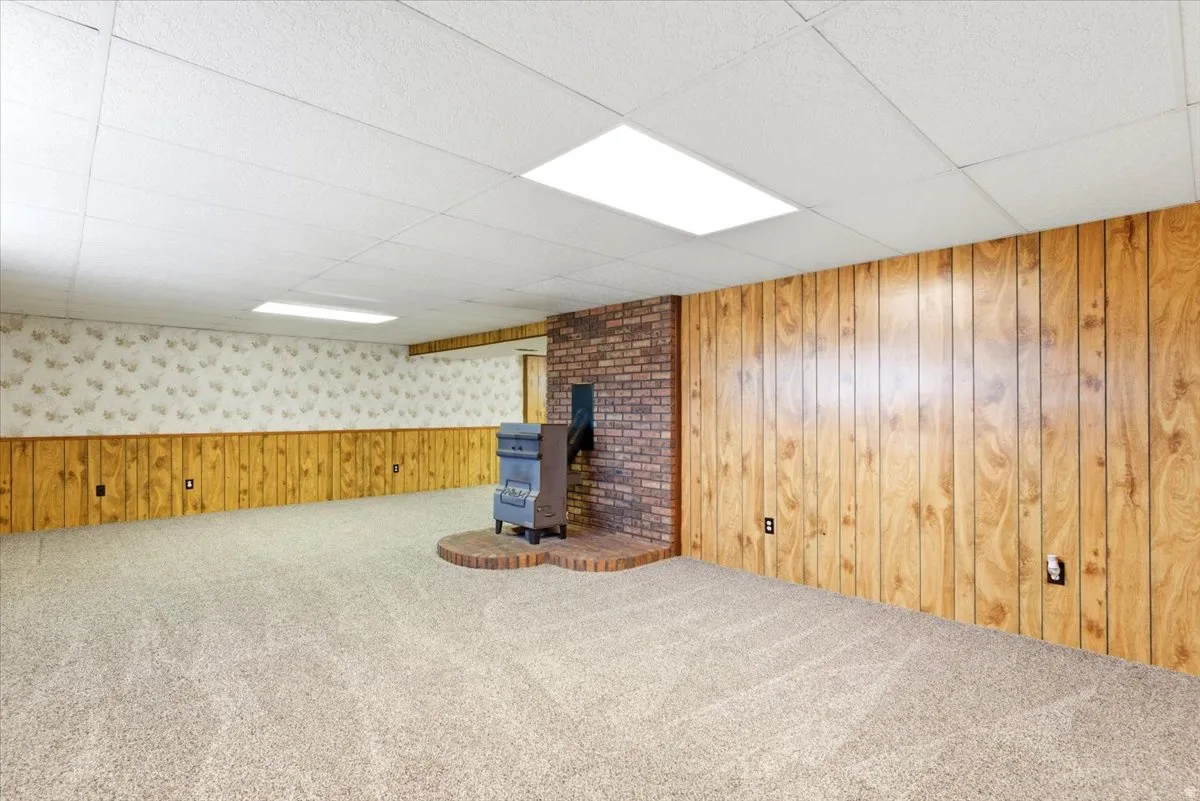 Spacious room featuring a brick wood-burning stove with a brick hearth, wall paneling, textured wallpaper, neutral carpeting, and drop ceiling with integrated lighting