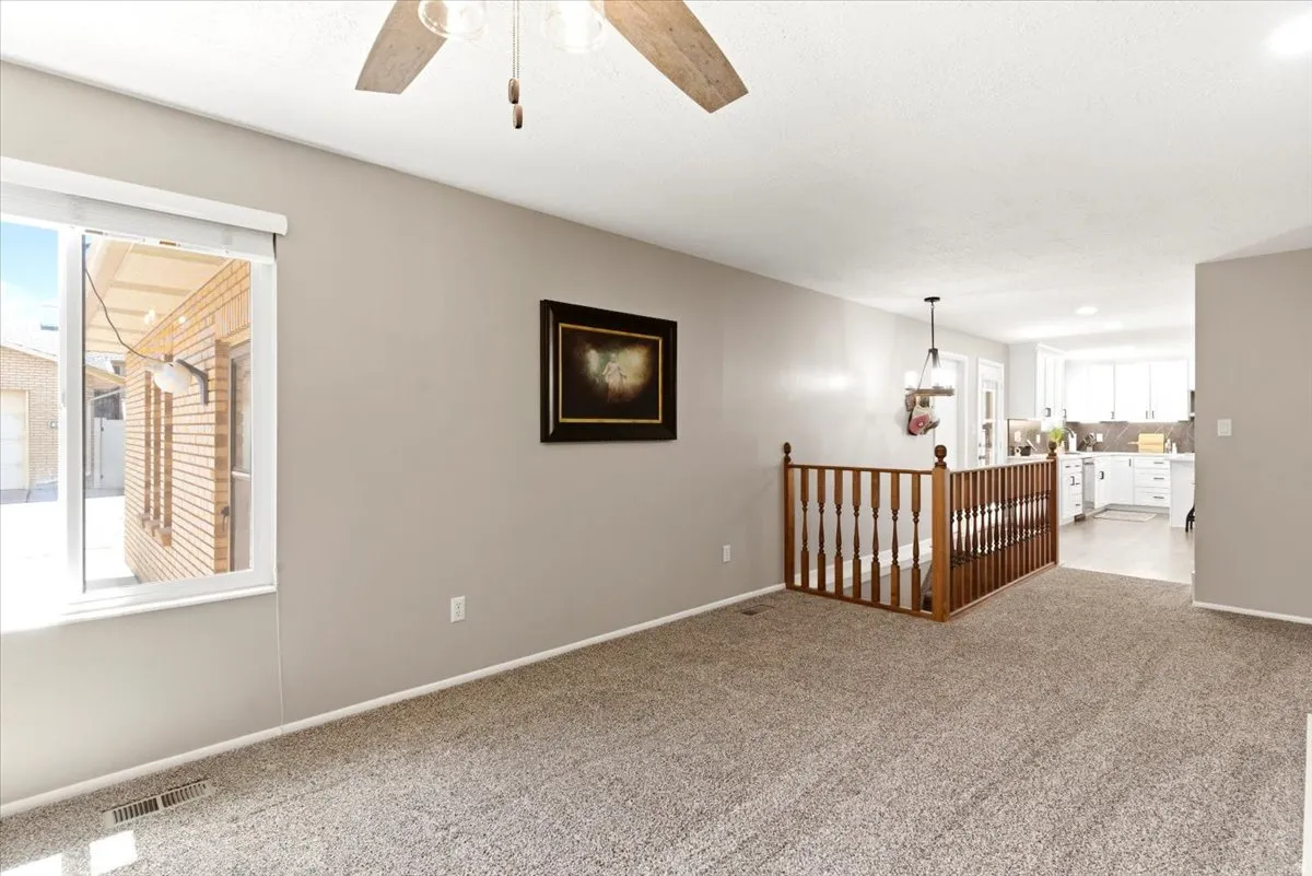 Spacious carpeted room featuring a ceiling fan, a large window, a wooden railing, and a partial view of a kitchen with white cabinetry