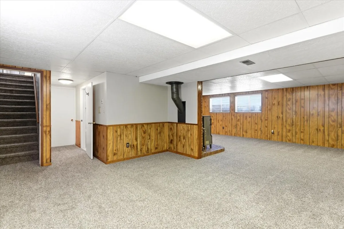 Spacious finished lower level featuring light gray carpet, wood-finish paneling, and a corner-set wood-burning stove with a brick hearth