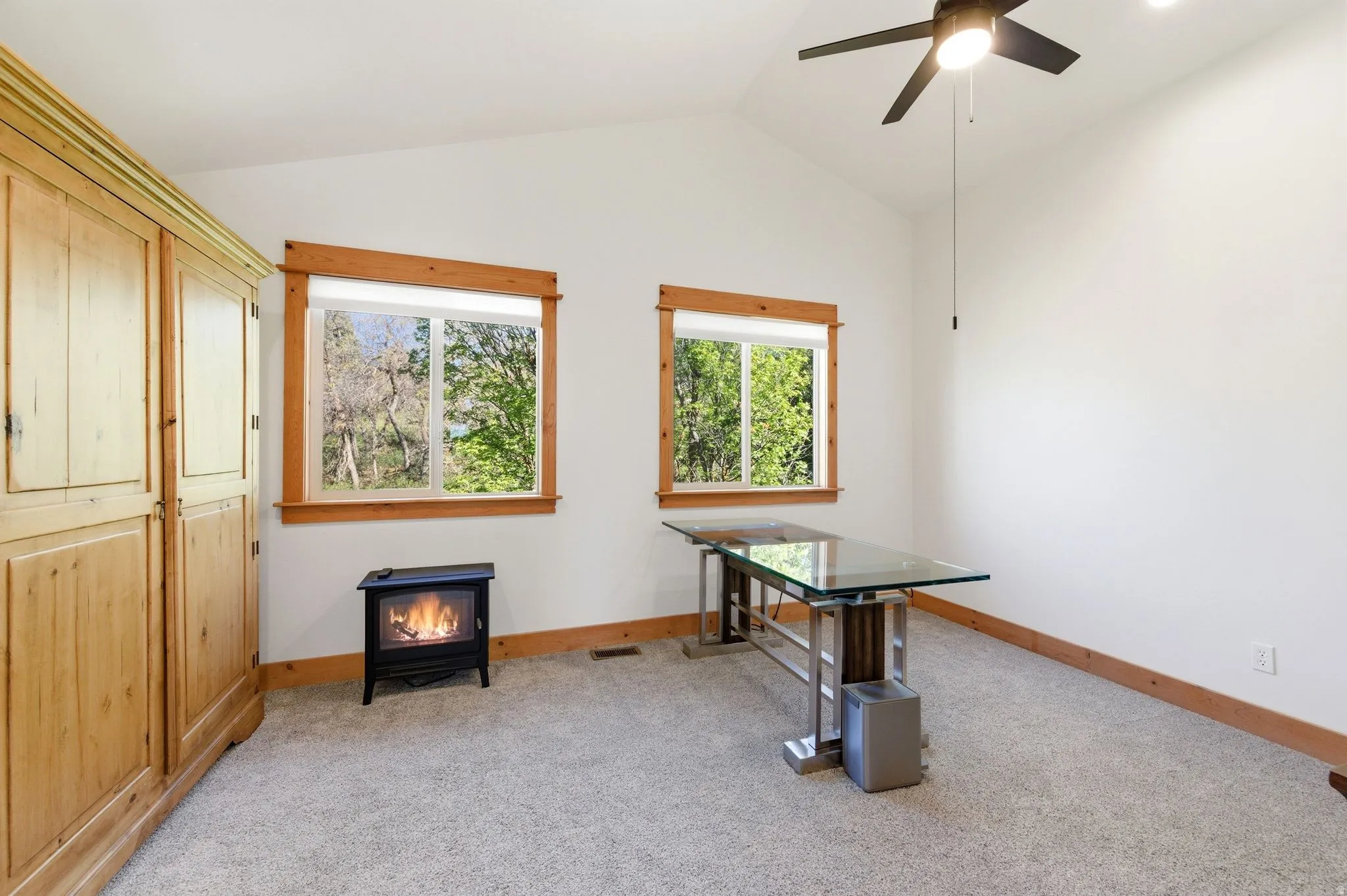 Vaulted ceilings with a ceiling fan, two double-hung windows with natural wood trim, wall-to-wall carpeting, and a freestanding fireplace