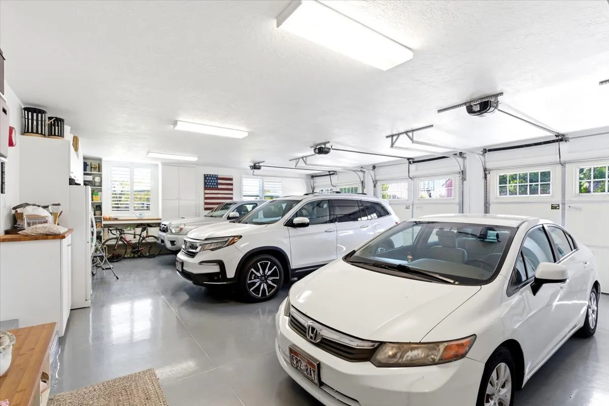 Three-car garage featuring segmented overhead doors with upper window panels, epoxy flooring, fluorescent ceiling lighting, and built-in white cabinetry with a wood-finish countertop