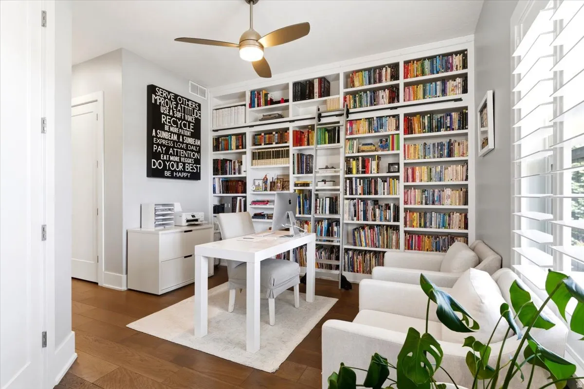 Dedicated office space featuring extensive built-in shelving, a rolling library ladder, wood-finish flooring, and a ceiling fan