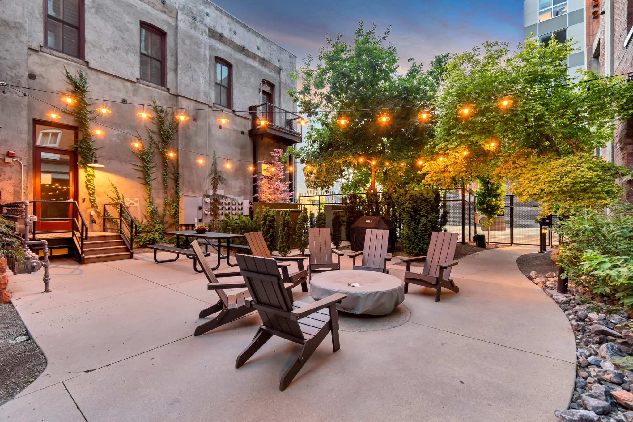 Spacious outdoor patio featuring a central concrete fire pit, string lighting, and extensive hardscaping