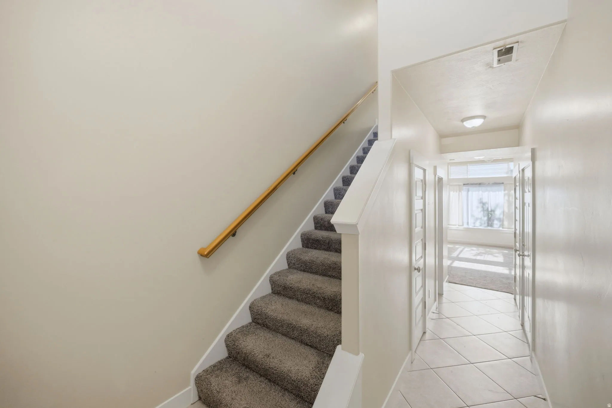 Carpeted staircase with a wood handrail, featuring a tiled hallway with recessed lighting and a bright room beyond