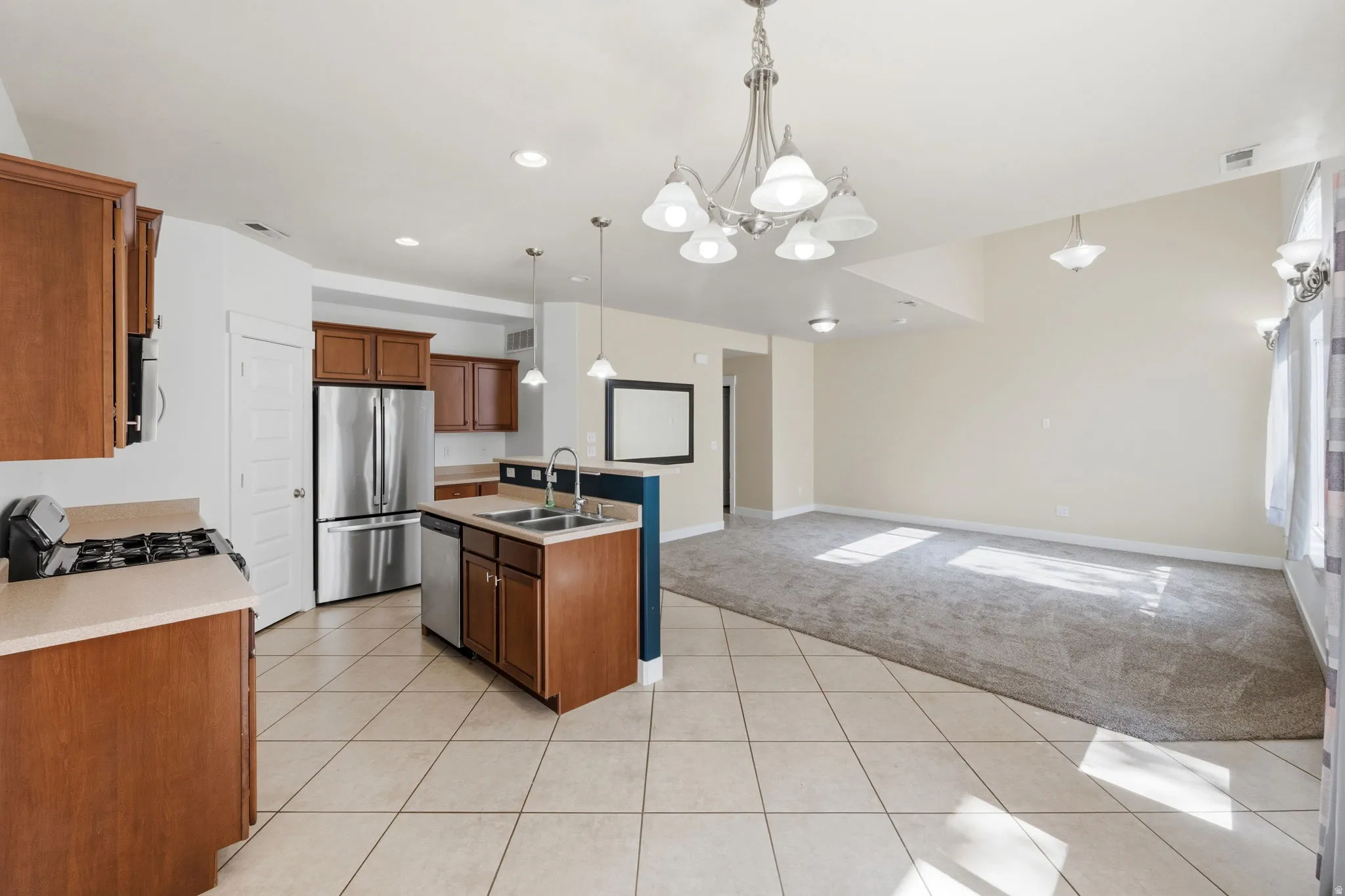 Open-concept kitchen and living area featuring stainless steel appliances, wood cabinetry, a central island with a double sink, tile flooring, and carpeted living space