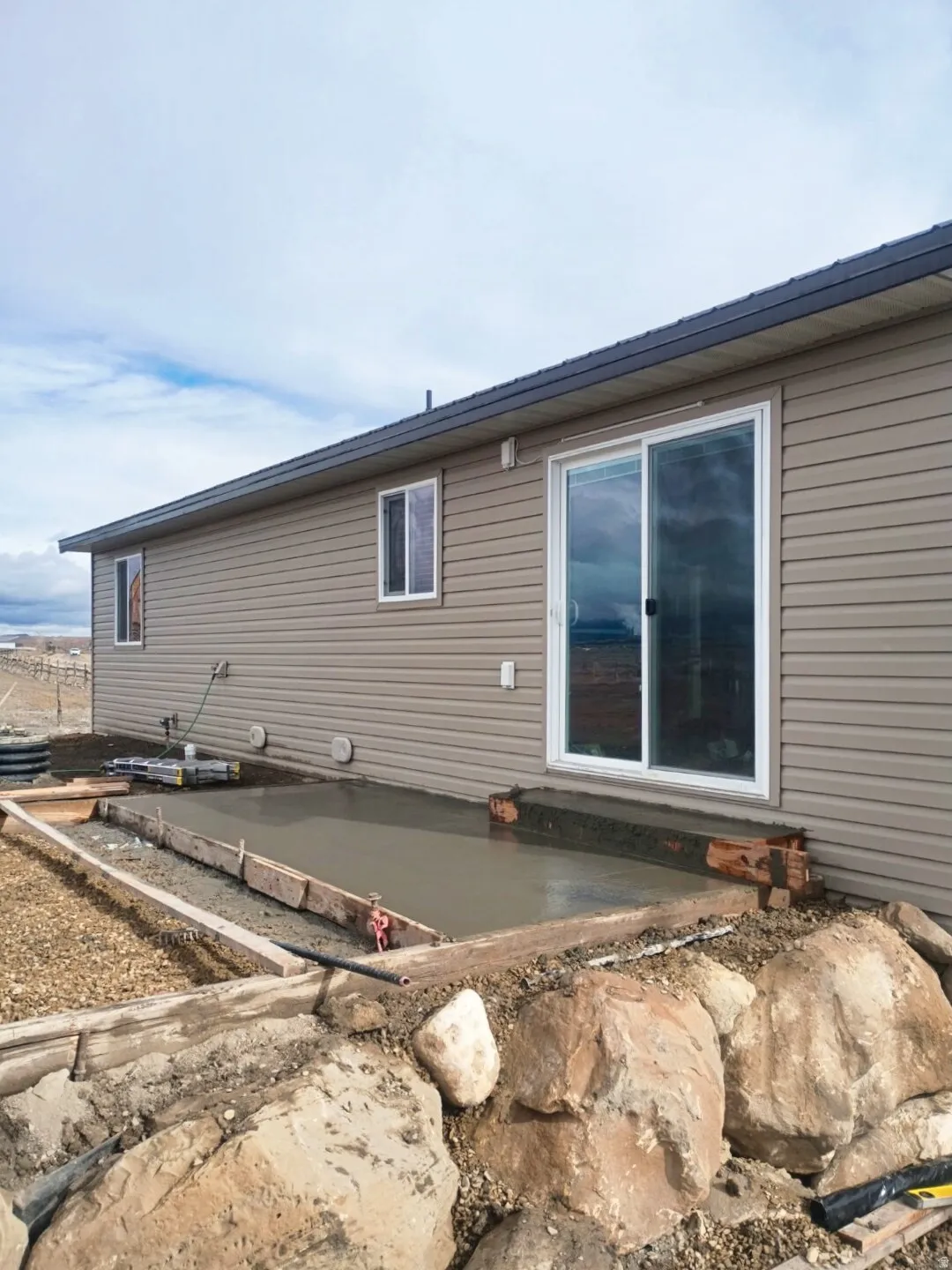 Exterior featuring tan horizontal siding, a sliding glass door, a newly poured concrete patio, and a rock retaining wall
