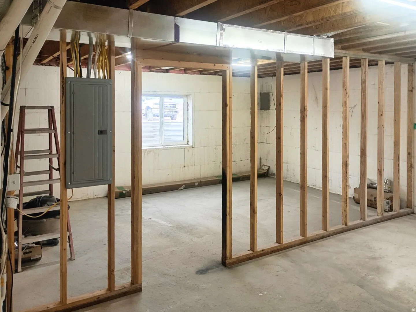 Unfinished basement area featuring framed partition walls, exposed overhead ductwork, and a visible electrical panel