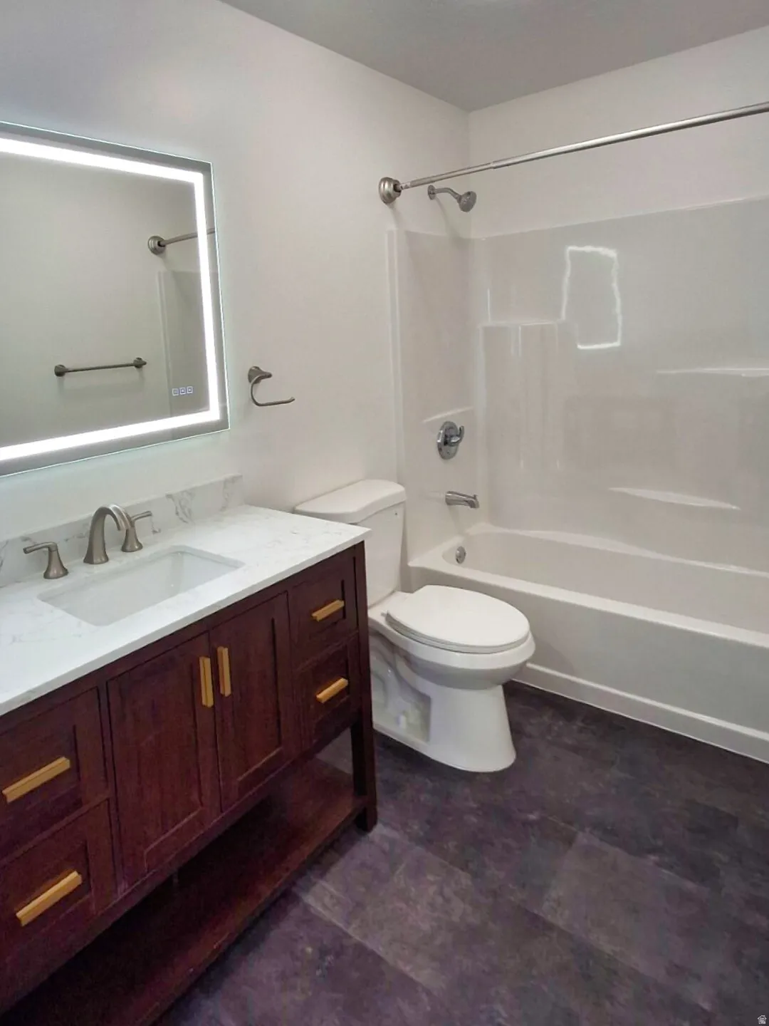 Bathroom featuring a dark wood-finish vanity with a white countertop and rectangular sink, an illuminated mirror, a white toilet, a white bathtub with a shower, and dark wood-finish flooring