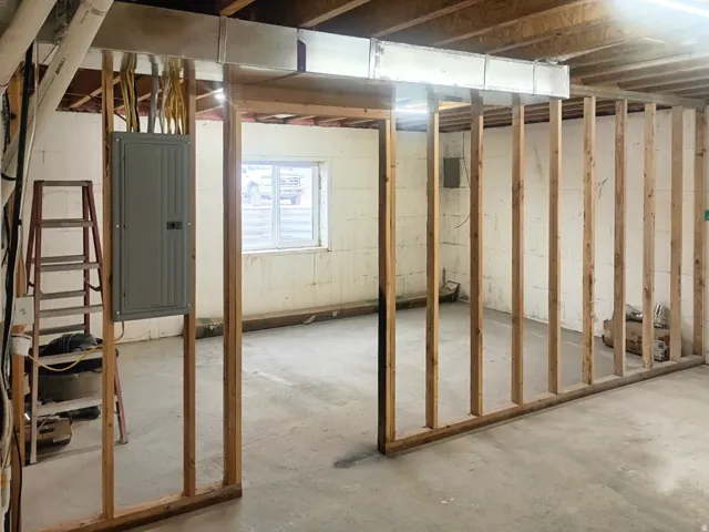 Unfinished basement area featuring framed partition walls, exposed overhead ductwork, and a visible electrical panel
