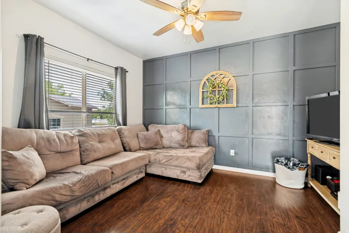 Interior space featuring a dark gray accent wall with geometric paneling, wood-finish flooring, a ceiling fan with light fixture, and a large window with horizontal blinds