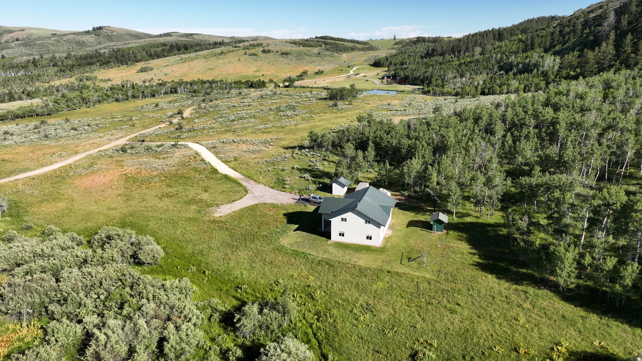 Expansive high-country setting featuring a white residence with a green roof, surrounded by mature trees, grasslands, and a dirt road