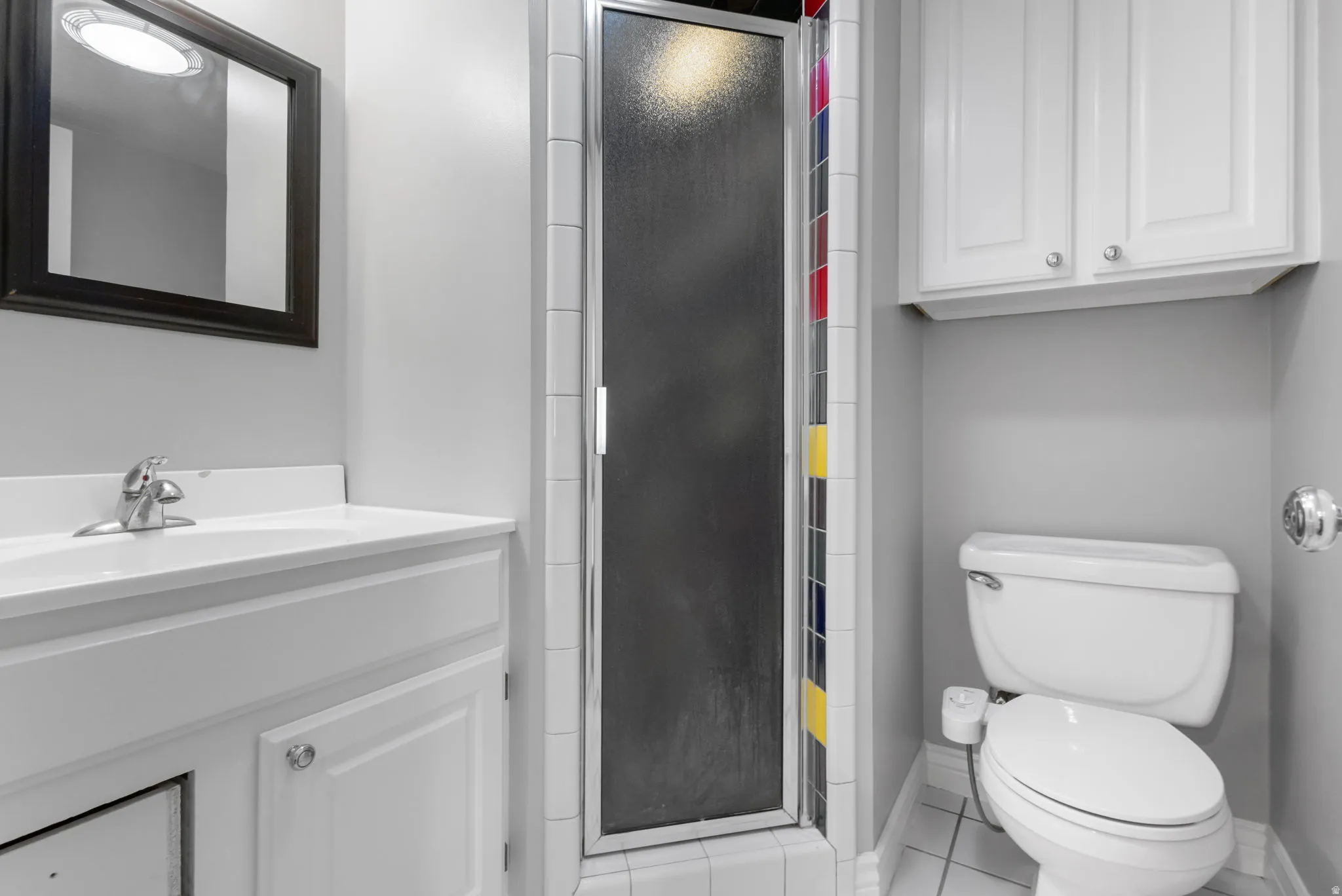 Three-piece bathroom featuring a shower with a frosted glass door, a toilet, a vanity with an integrated sink, a framed mirror, and upper cabinetry