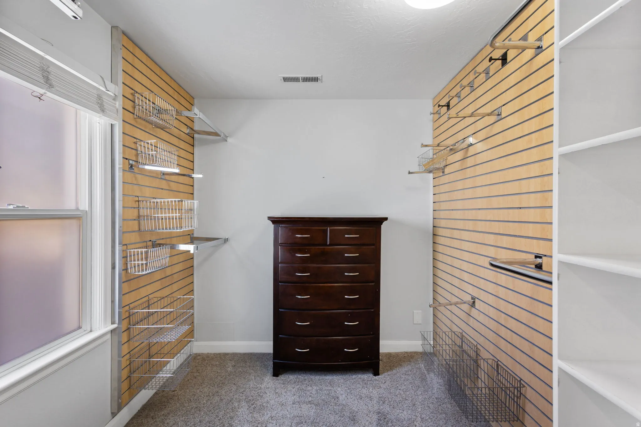 Customizable closet system featuring slatwall panels with adjustable shelving and wire baskets, built-in white shelving, and a double-hung window with privacy film