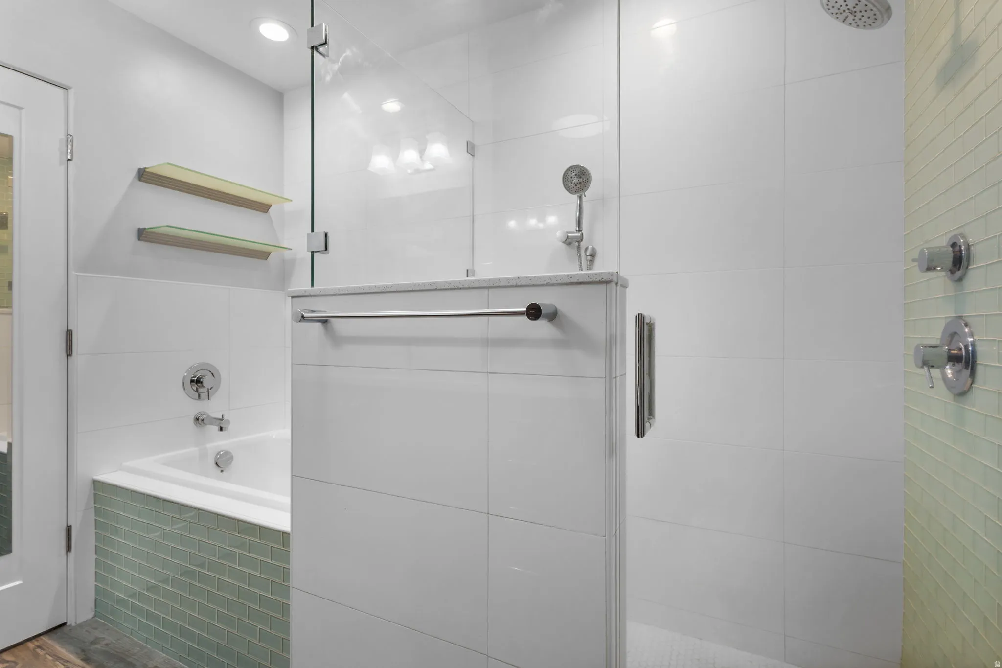 Glass-enclosed shower with two showerheads alongside a built-in tub featuring light green glass tile accent and white tile surround and chrome fixtures