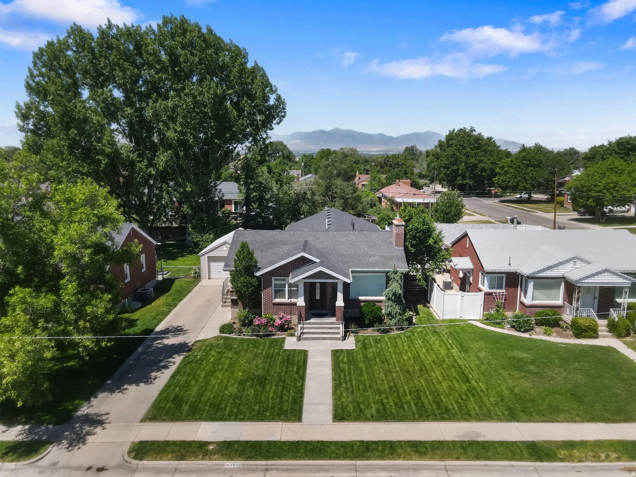 Brick exterior home featuring a well-maintained lawn, mature landscaping, an attached garage, a paved driveway, and established trees
