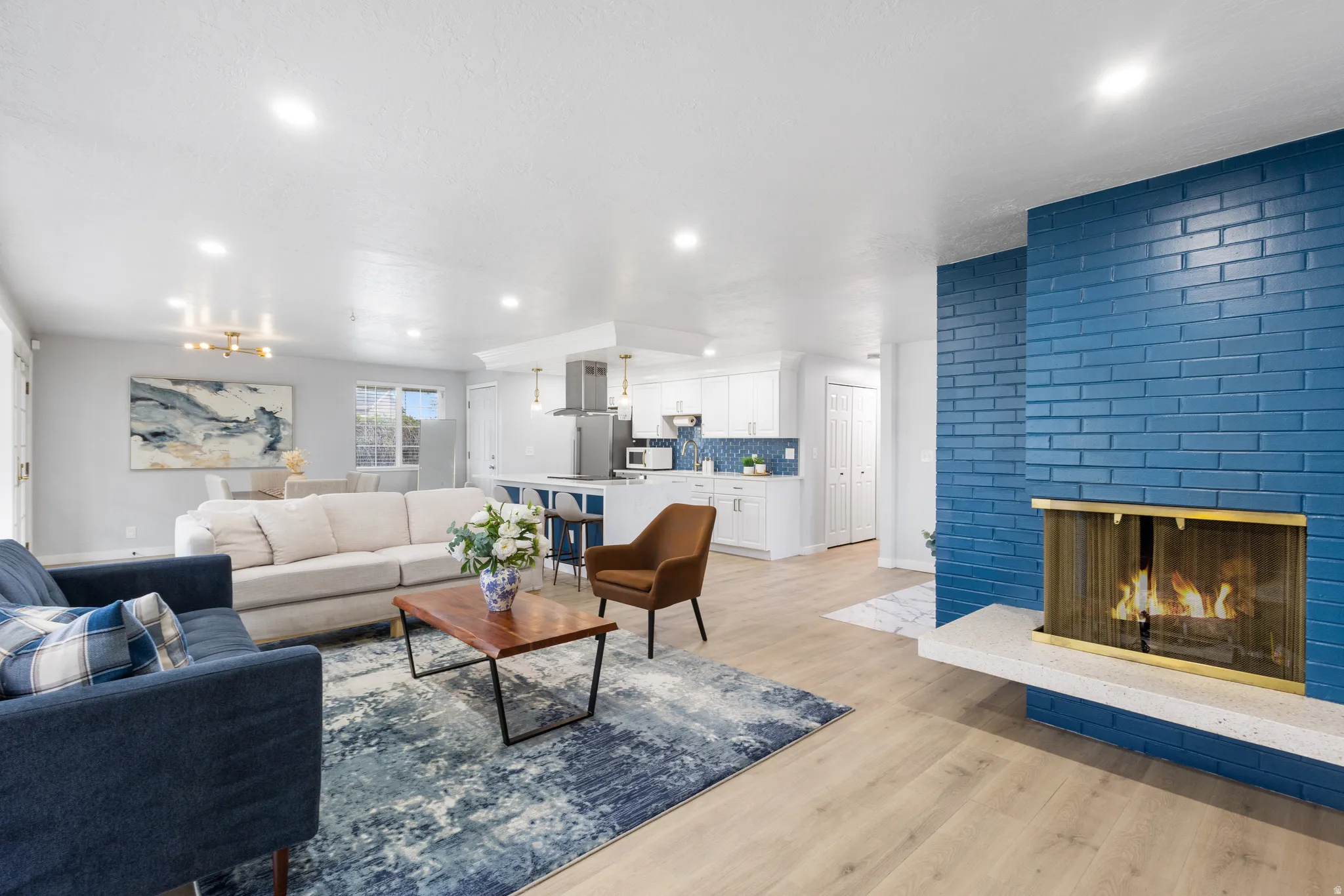 Living space featuring a prominent blue brick fireplace with a white hearth, light wood-finish flooring, recessed lighting, and a kitchen with white cabinetry and stainless steel appliances