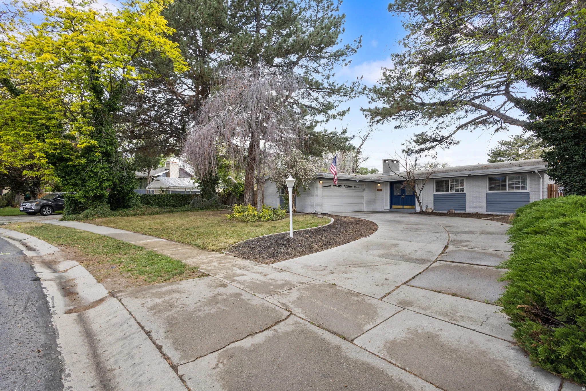 Ranch-style residence featuring a curved concrete driveway, mature trees, an attached garage, and a charcoal chimney