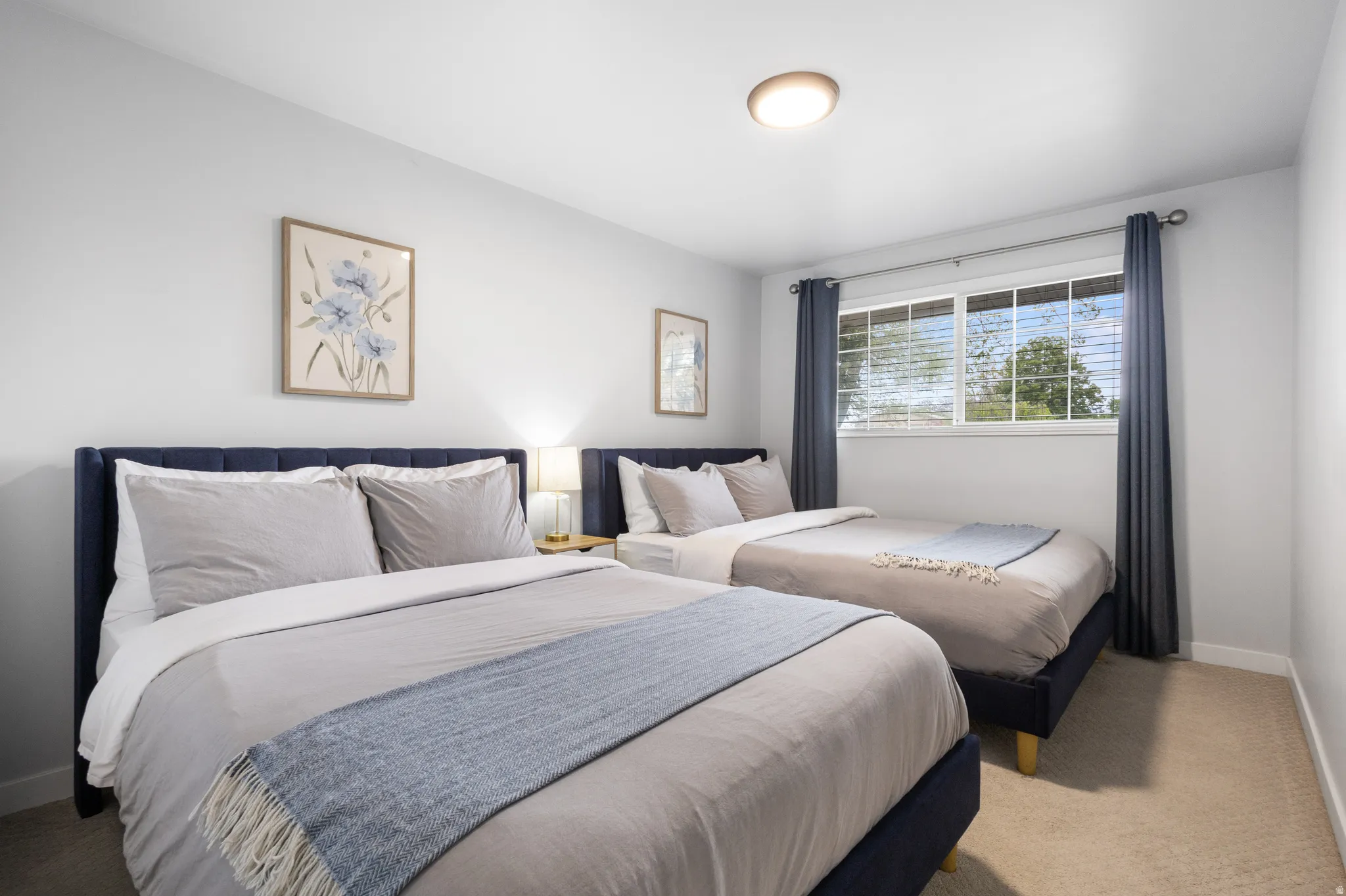 Bedroom featuring two bed frames with upholstered headboards, a window with blinds and dark curtains, and light-colored carpeting