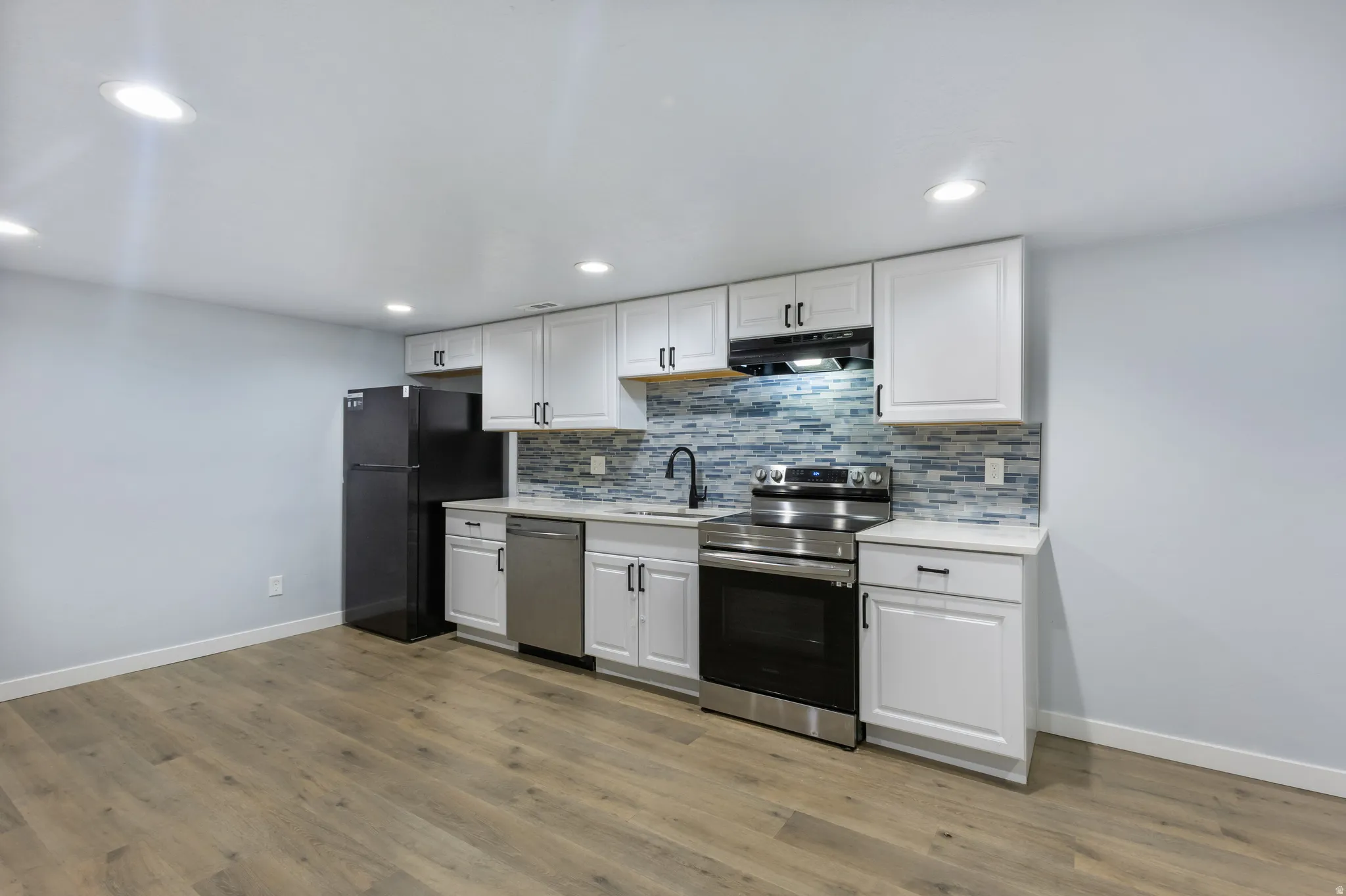 Kitchenette featuring white cabinetry with dark hardware, a mosaic tile backsplash, stainless steel appliances, and wood-finish flooring