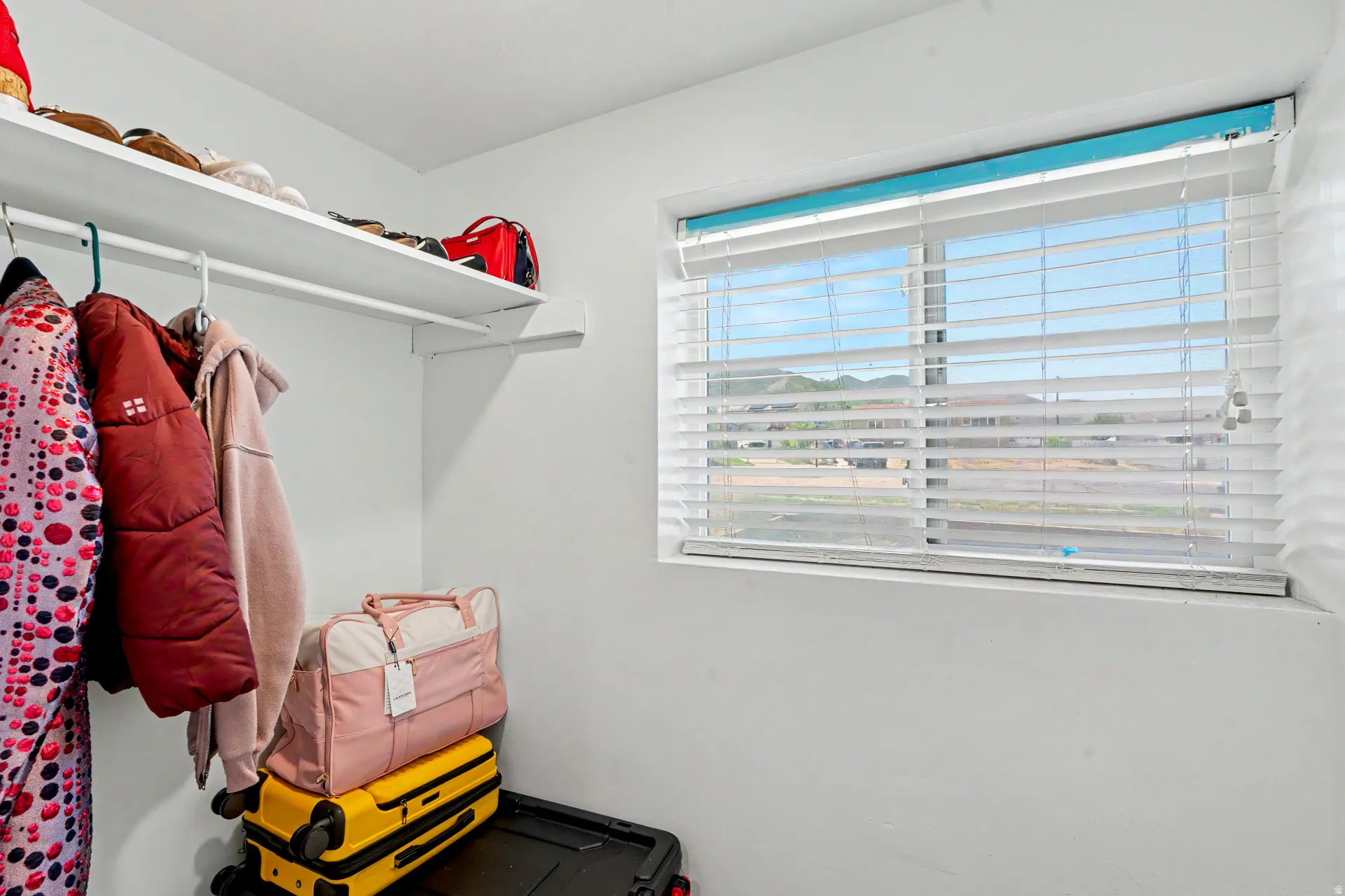 Coat Closet space featuring a window with white horizontal blinds, a white wall-mounted shelf, and a white hanging rod